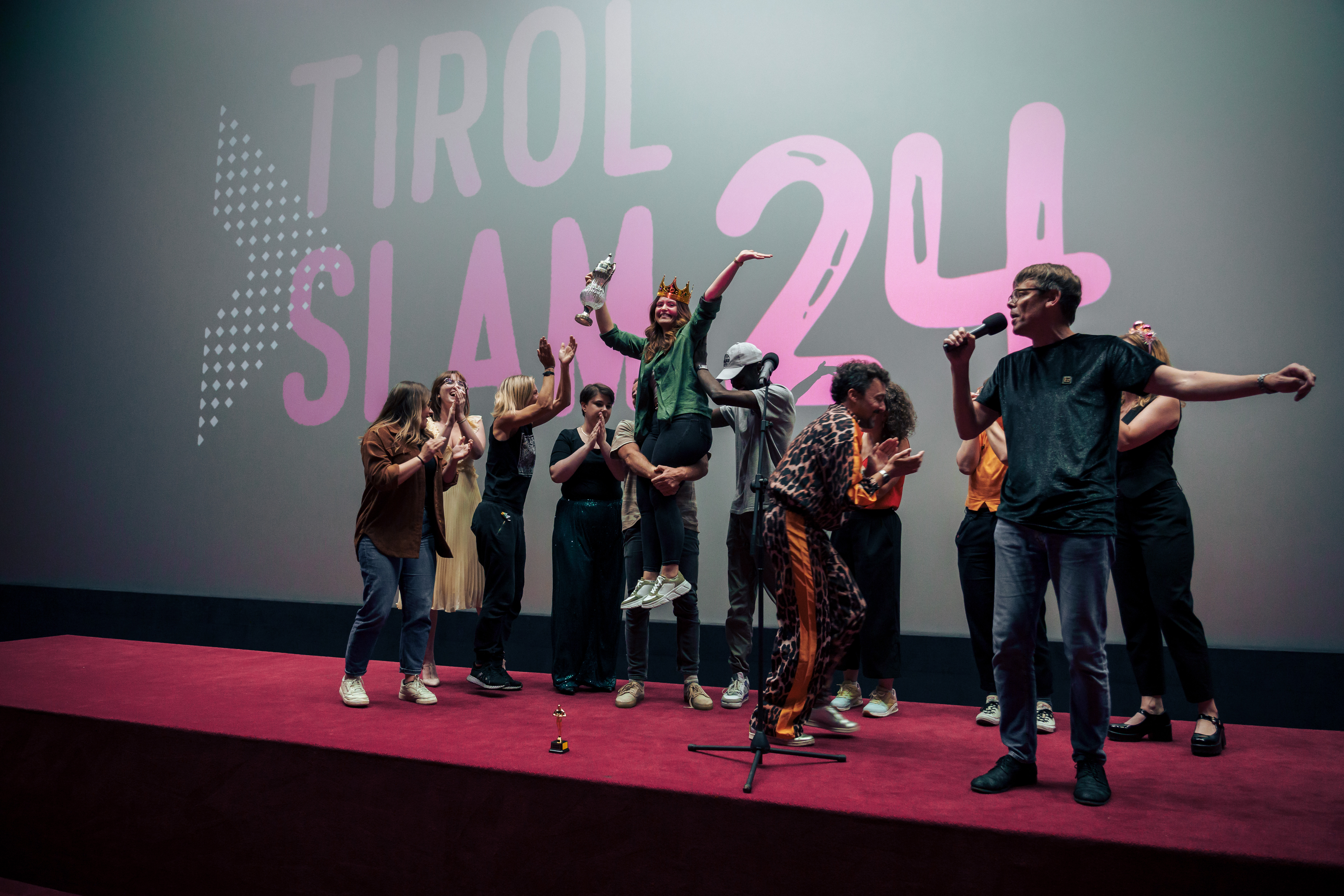 12 jubelnde Menschen auf einer roten Bühne vor einer Leinwand auf der "Tirol Slam 24" steht. Eine Person mit Krone und Pokal wir hochgehoben.