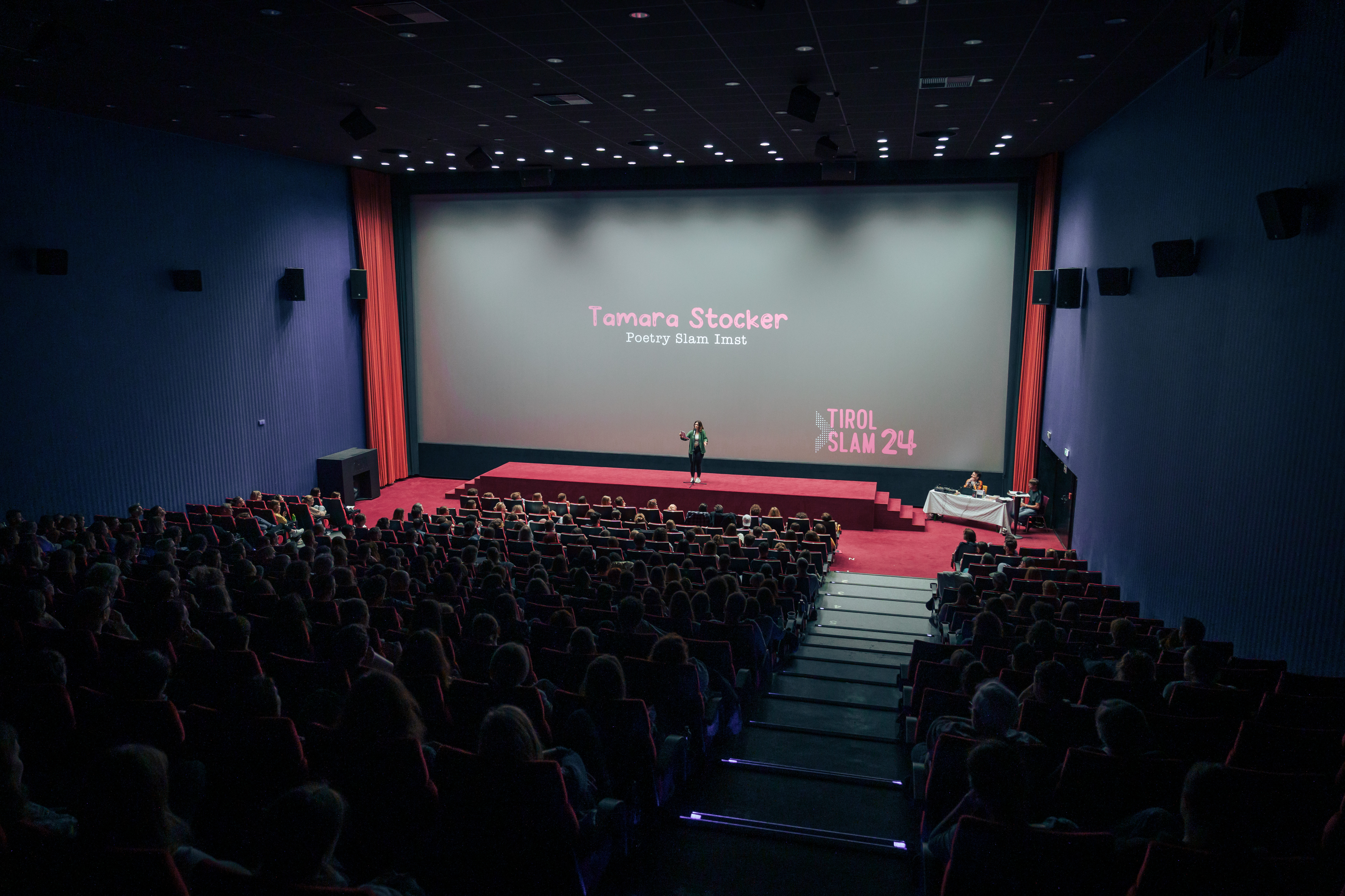 In einem großen Kinosaal steht eine Person auf der Bühne. Im Hintergrund auf der Leinwand steht: "Tamara Stocker - Poetry Slam Imst" und "Tirol Slam 24".