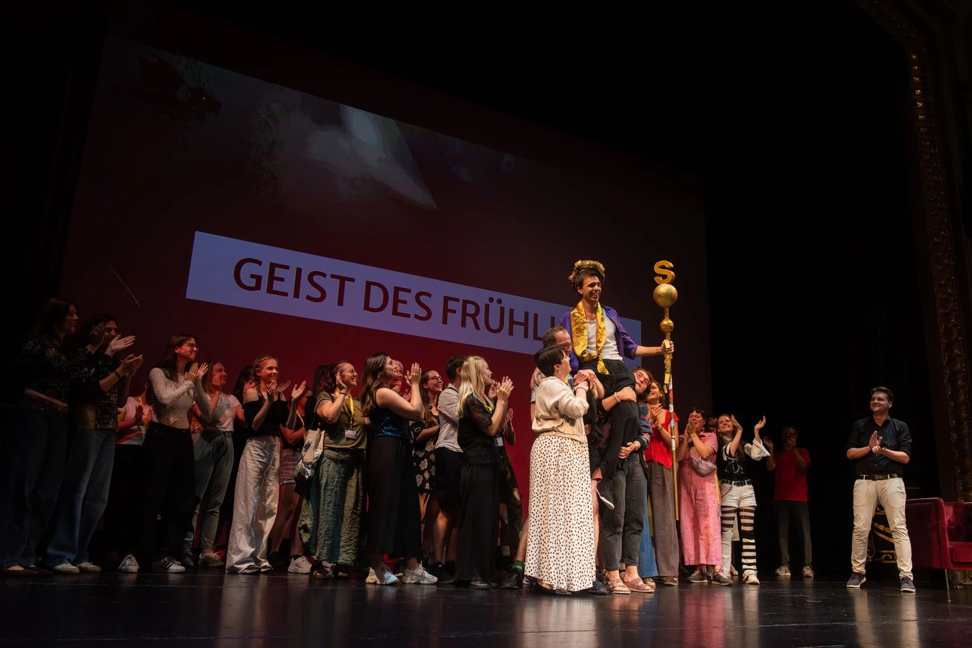 Eine Person mit Scherpe, Krone und Szepter wird von einigen Personen auf einer Bühne auf den Schultern getragen, im Hintergrund stehen noch mehr Menschen und auf der Leinwand groß "Geist des Frühlings"